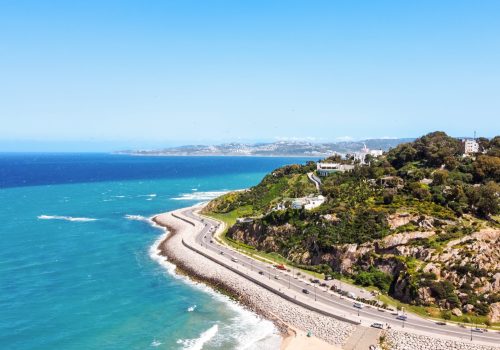 Aerial drone view of Tangier, Morocco. Strait of Gibraltar coastline, rocky hills with residential buildings and greenery, highway with moving cars